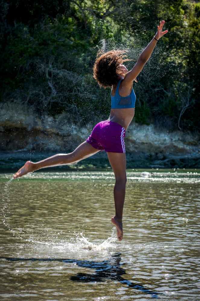 woman in blue sports bra and purple shorts leaping above body of water